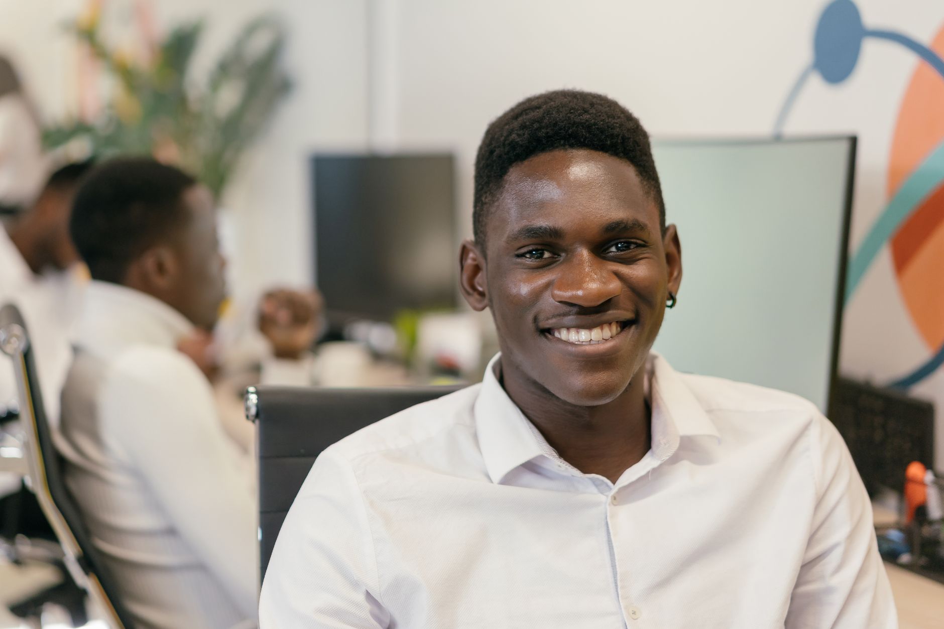 Jovem sorridente de camisa branca à mesa de trabalho num ambiente de escritório moderno.