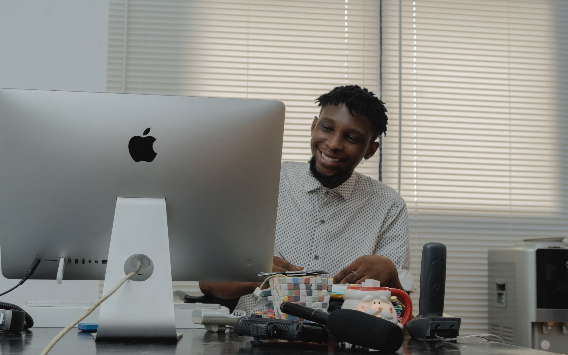Homem africano sorridente a trabalhar à mesa com um computador num espaço de escritório bem iluminado.