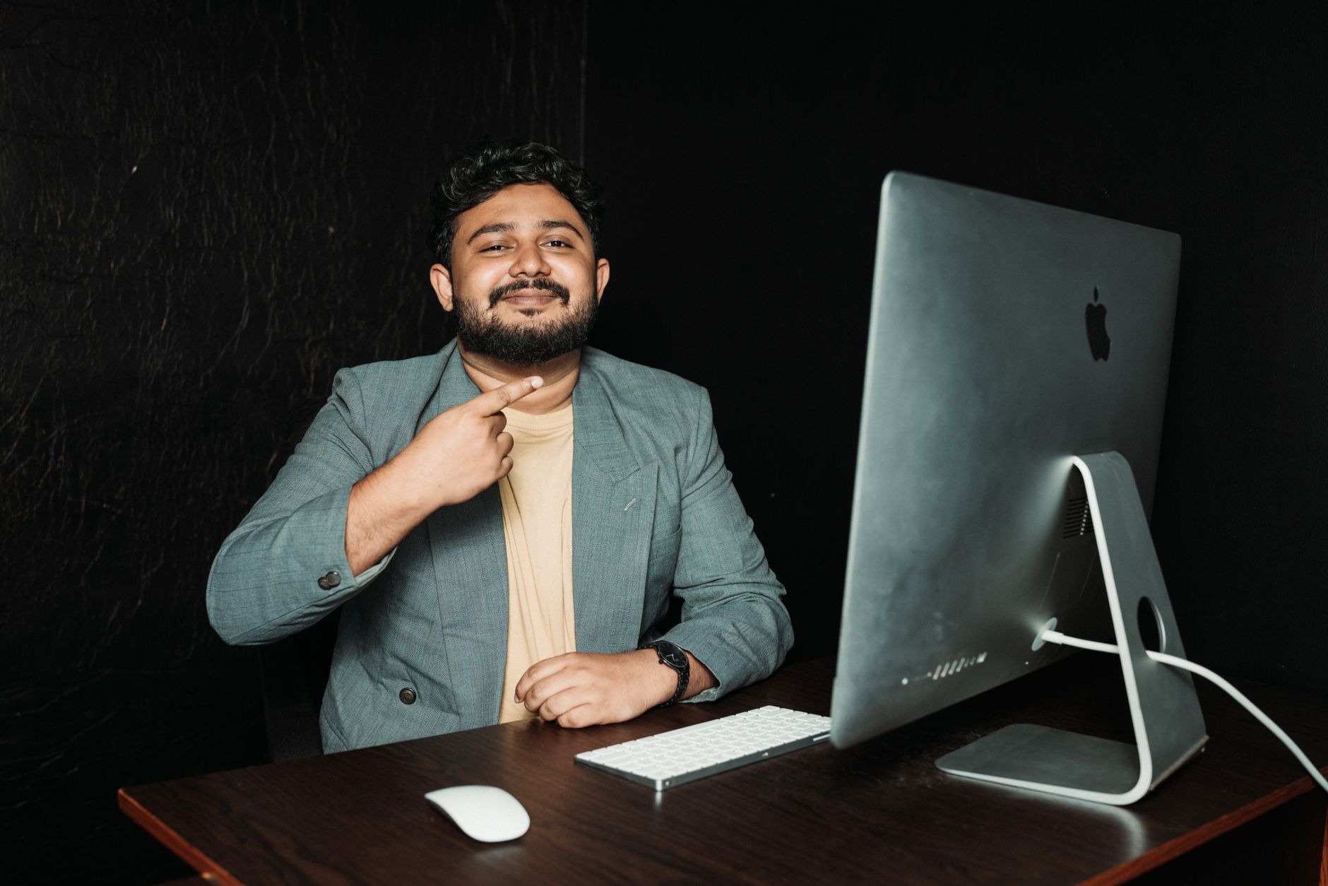 Homem sorridente num ambiente de escritório moderno, junto a um computador de mesa, num contexto profissional.