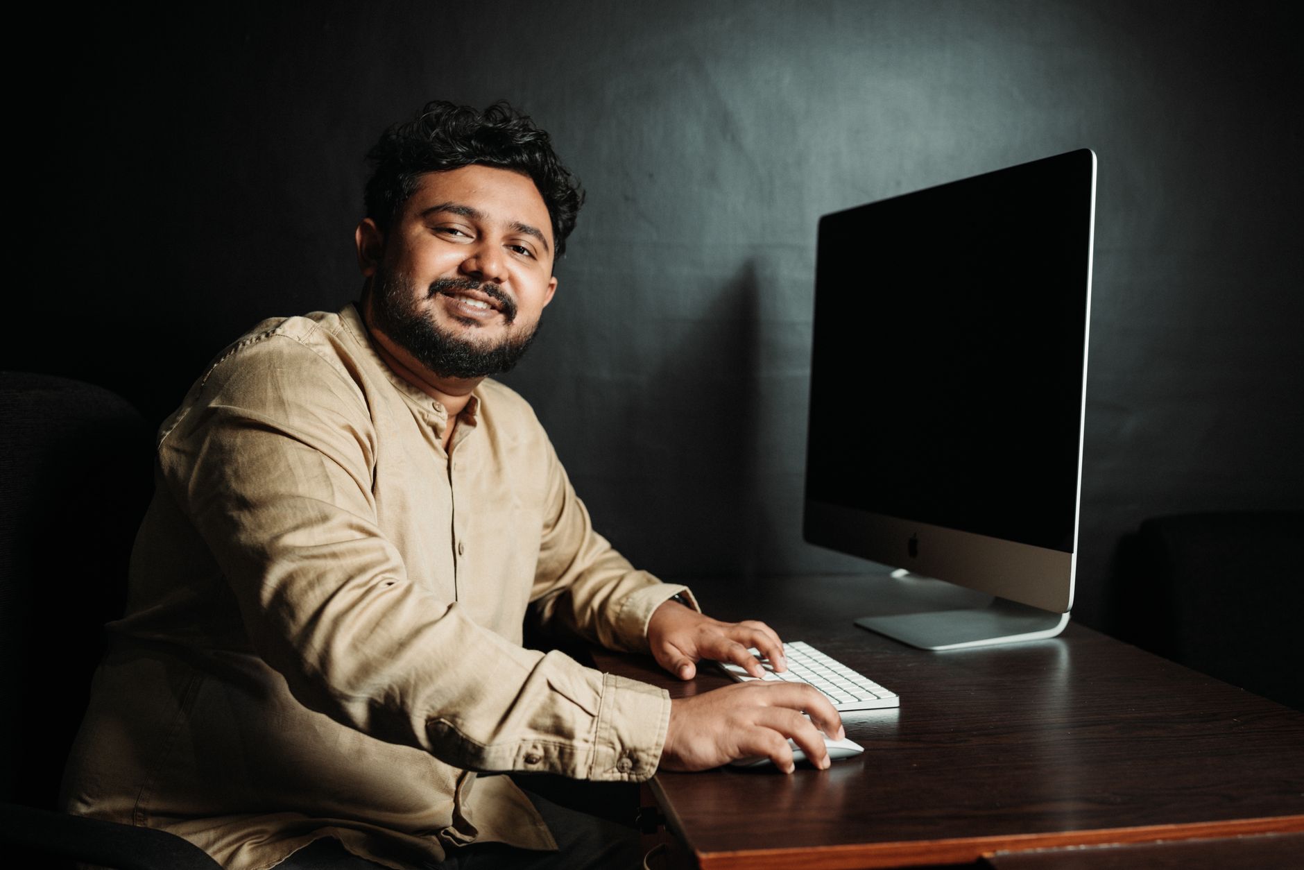 Homem sorridente em ambiente casual a trabalhar num computador de mesa, num escritório com pouca luz.
